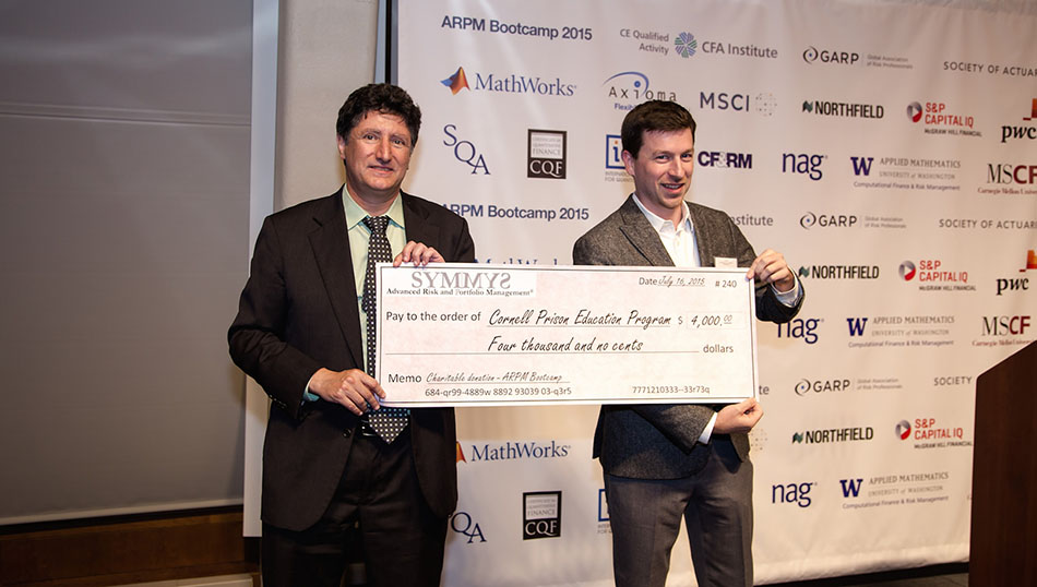 Two men hold a large $4,000 ceremonial check from SYMMYS Advanced Risk and Portfolio
                         Management to a representative for the Cornell Prison Education Program. The background is a
                         sponsor banner for the event.